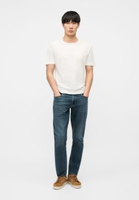 Jeune homme debout, les mains dans les poches, portant une chemise blanche à manches courtes, un jean bleu et des baskets marron, sur fond blanc uni.
