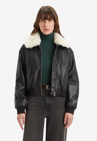 Čierna skrátená kožená bomber bunda s huňatým bielym golierom, zapínaním na zips a rebrovanými manžetami, nosená cez zelený rolák.