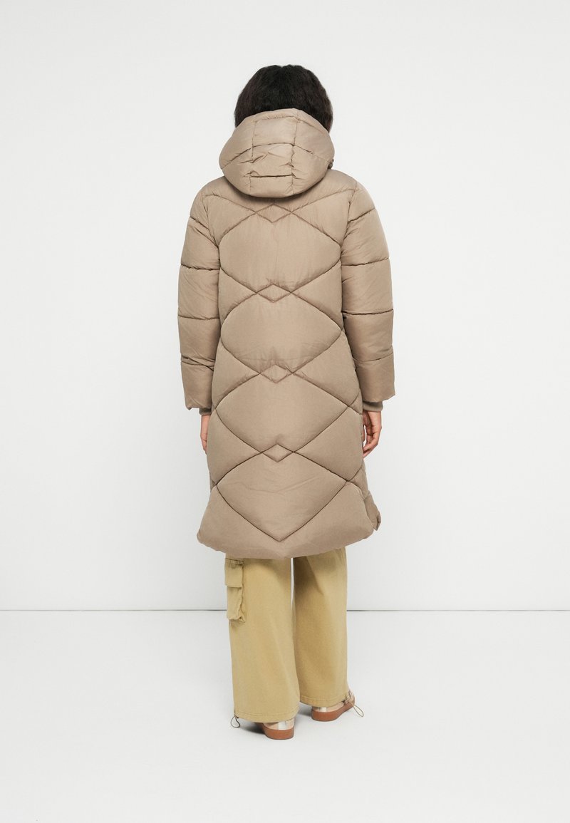 Personne portant un long manteau matelassé beige avec capuche, un large pantalon beige et des chaussures marron, se tenant contre un fond blanc, dos tourné vers le spectateur.