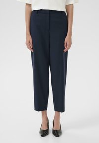 Pantalons tapering navy avec une texture lisse, dotés d'une taille et d'un pli devant repassé, assortis à des escarpins noirs à talons pointus.