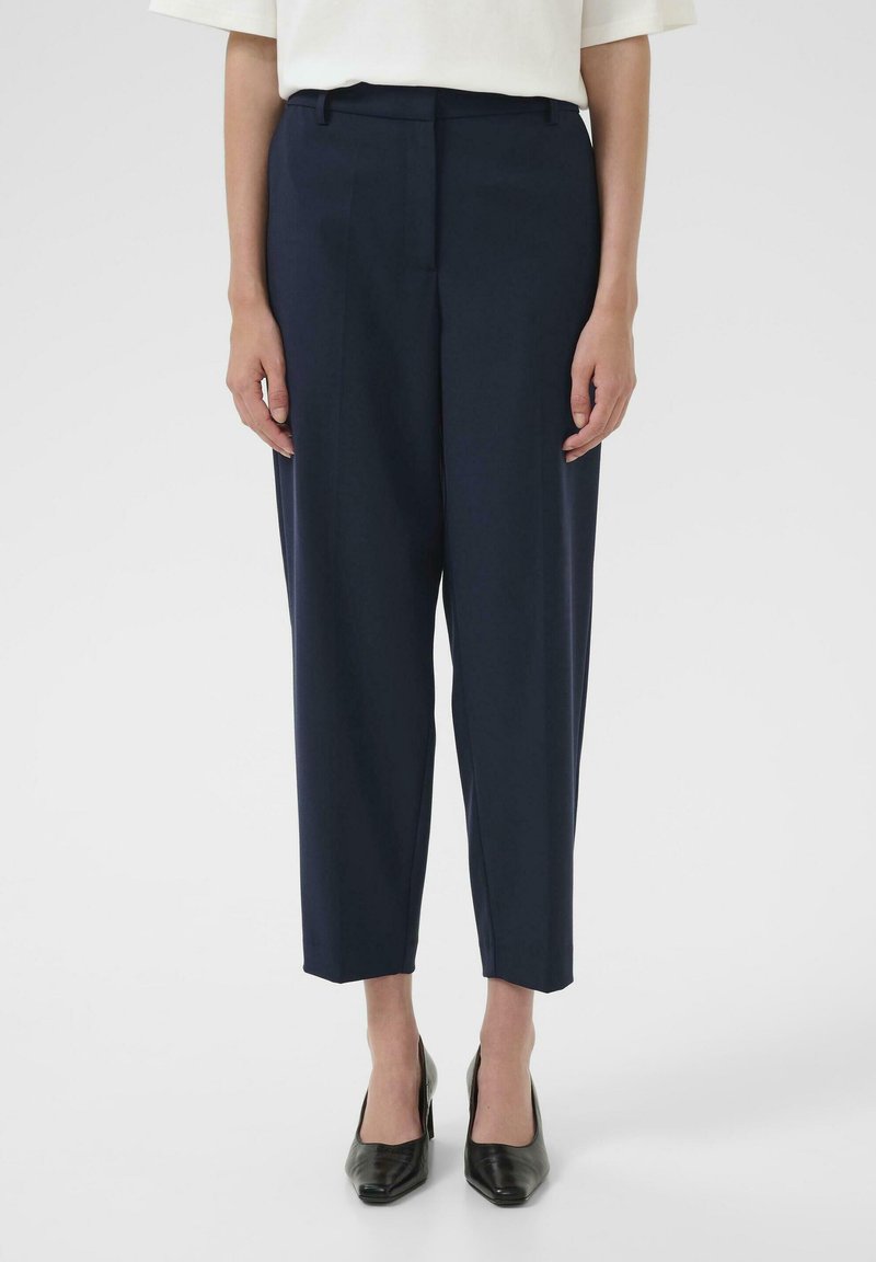 Pantalons tapering navy avec une texture lisse, dotés d'une taille et d'un pli devant repassé, assortis à des escarpins noirs à talons pointus.