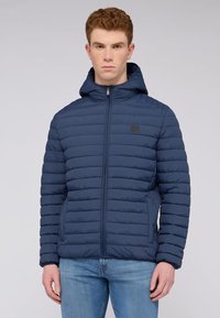 Námořnická puffer bunda se zipem vpředu, horizontálně prošívaným designem, nastavitelnou kapucí a bočními kapsami. Tkanina vypadá hladce se mírným leskem.