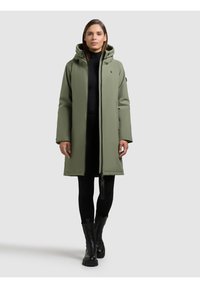 Lungo cappotto imbottito verde oliva con cappuccio, chiusura con cerniera, tasche laterali e una finitura opaca elegante. Abbinato a pantaloni neri e stivali.