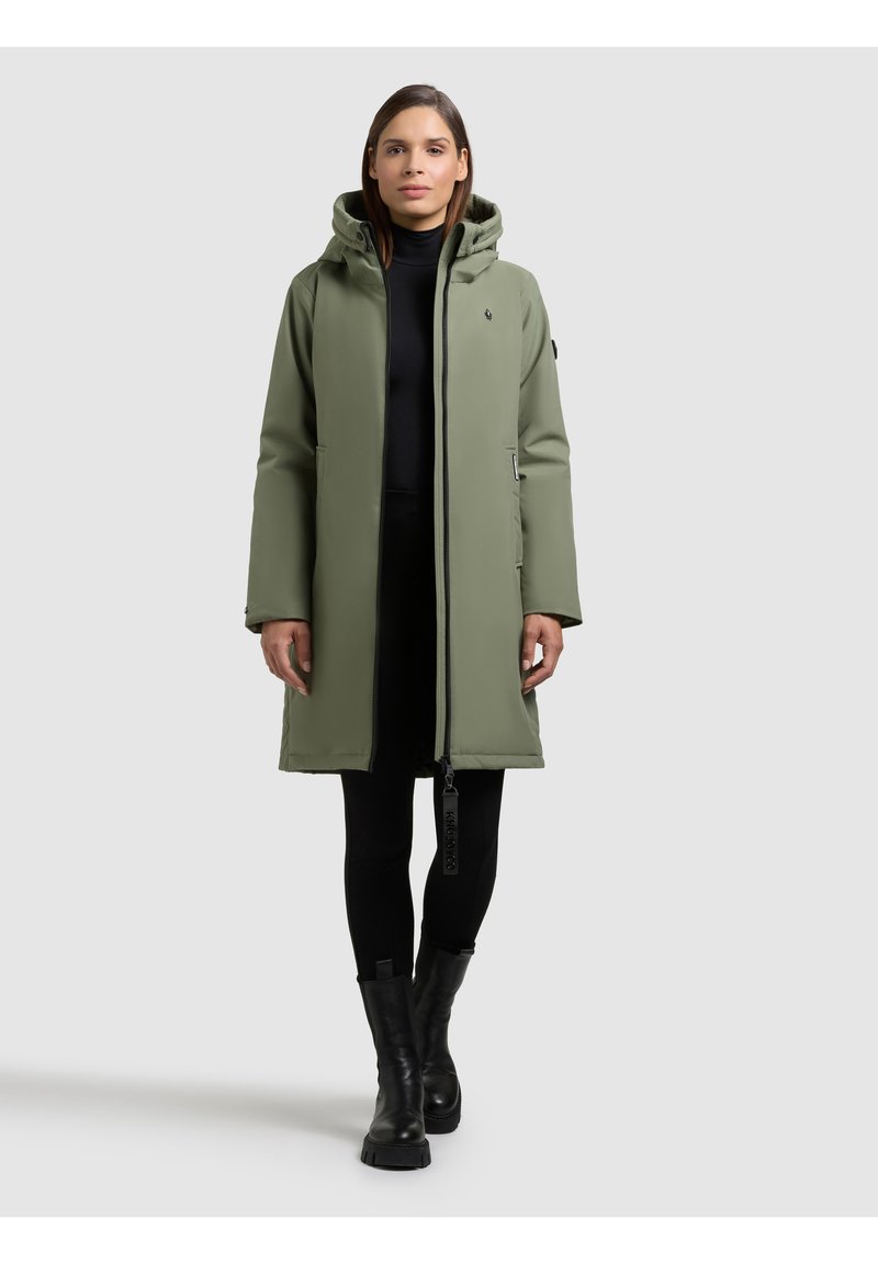 Lungo cappotto imbottito verde oliva con cappuccio, chiusura con cerniera, tasche laterali e una finitura opaca elegante. Abbinato a pantaloni neri e stivali.
