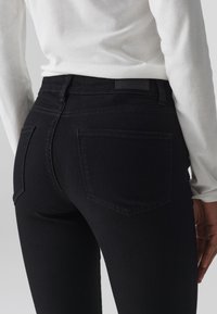 Jeans negros de talle alto con un corte ajustado, con un diseño de cinco bolsillos y sutiles detalles de costura, combinados con una camisa blanca de manga larga.
