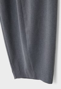 Gros plan sur la partie inférieure d’un pantalon gris foncé sur mesure montrant la texture du tissu et les détails des plis sur un fond uni.