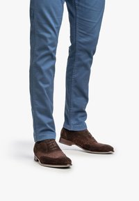 Hombre vestido con pantalones azules y zapatos de vestir de ante marrón con cordones, de pie sobre un fondo blanco.