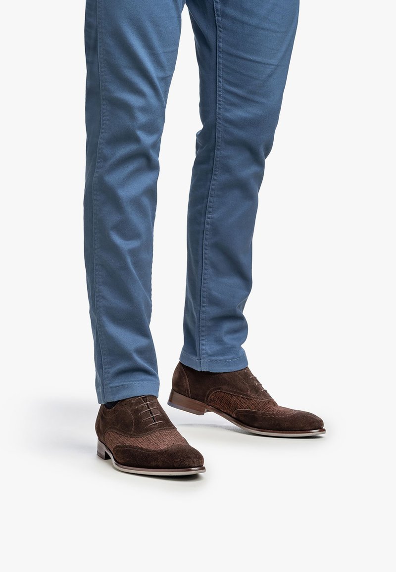 Hombre vestido con pantalones azules y zapatos de vestir de ante marrón con cordones, de pie sobre un fondo blanco.