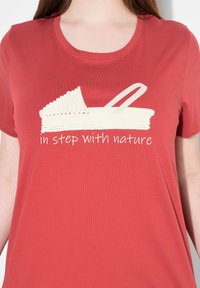 Camiseta de algodón roja con un gráfico de una cesta tejida y el texto "en sintonía con la naturaleza" en una fuente cursiva blanca en la parte frontal.