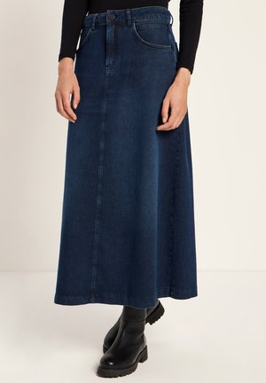 Frau in einem dunkelblauen Jeansrock mit vorderen Nähten, schwarzem langärmeligen Top und schwarzen Lederstiefeletten vor einem hellen Hintergrund.