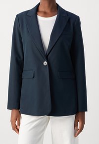 Námořnický blazer s jedním knoflíkem, dvěma předními kapsami, klopami s výstřihem, přiléhavým střihem a hladkým materiálem, v kombinaci s bílým topem a světlými kalhotami.