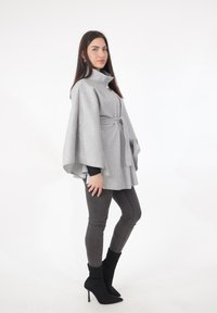 Grijze wollen cape met hoge kraag, tailleband en wijde mouwen, gecombineerd met strakke donkere jeans en zwarte enkellaarsjes met puntige neus.