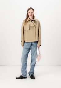 Giacca corta beige con collo oversize e bottoni neri; abbinata a jeans azzurri a gamba larga e una piccola borsa rosa. Mocassini neri completano il look.
