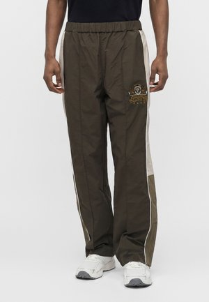 Homme portant un pantalon de survêtement vert olive avec des bandes blanches sur les côtés, logo brodé sur la cuisse gauche, et des baskets blanches, debout devant un fond uni.