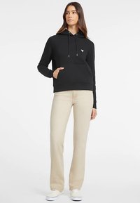 Zwarte hoodie met een voorzak, verstelbare trekkoorden en een klein logo op de borst, gecombineerd met licht beige broek en witte schoenen.