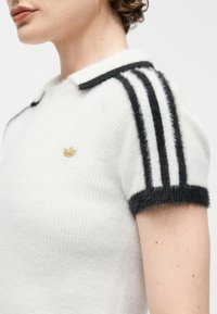 Κοντινό πλάνο ενός ατόμου που φοράει λευκό, αφράτο κοντομάνικο τοπ με μαύρες ρίγες στα μανίκια και χρυσό λογότυπο Adidas στο στήθος.