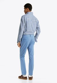 Chemise à carreaux bleus et blancs, manches longues, vue de dos, associée à un pantalon bleu clair et des baskets brunes avec des accents blancs.