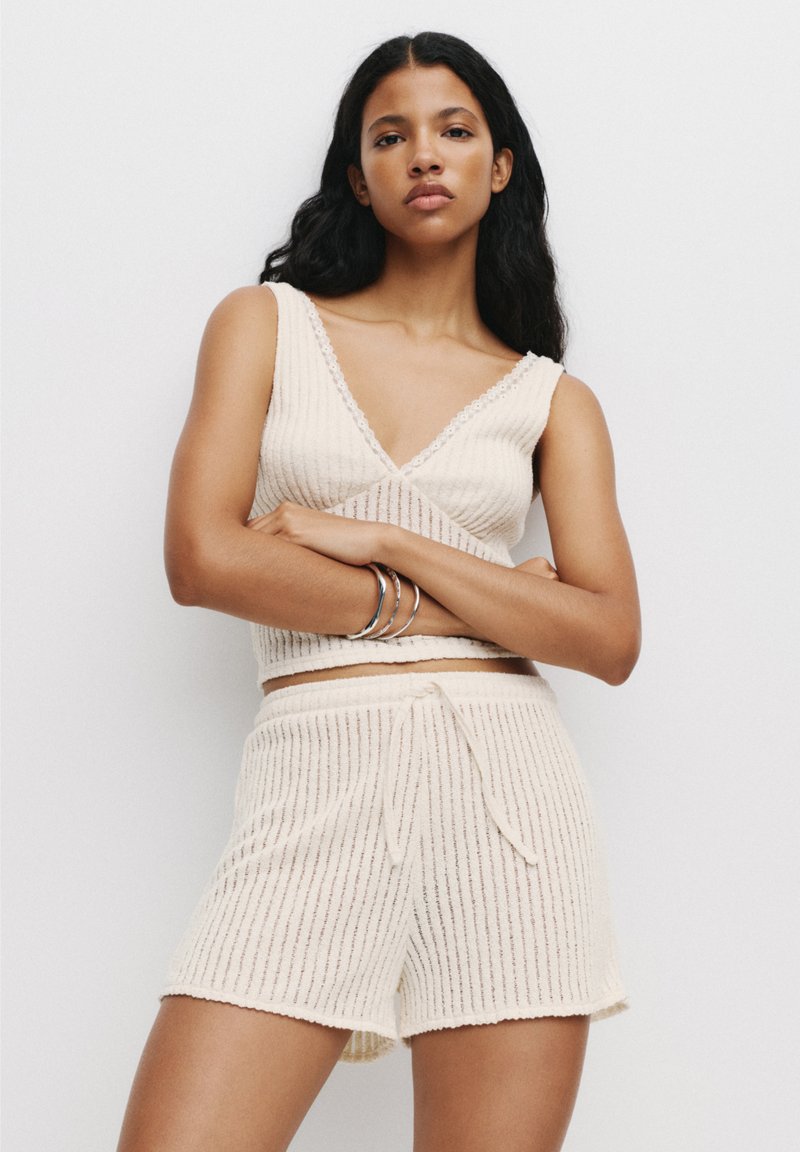 Crème-kleurige gebreide loungewear set met een mouwloze top met diepe V-hals en shorts met trekkoord, voorzien van verticale geribbelde textuur en accentrandjes.