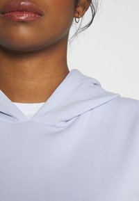 Sweat à capuche bleu clair à texture douce, avec une poche avant et une capuche à cordon, porté par-dessus un t-shirt blanc.