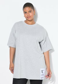 Camiseta de gran tamaño en gris claro con mangas cortas, escote redondo y una etiqueta lateral con texto negro sobre fondo blanco.
