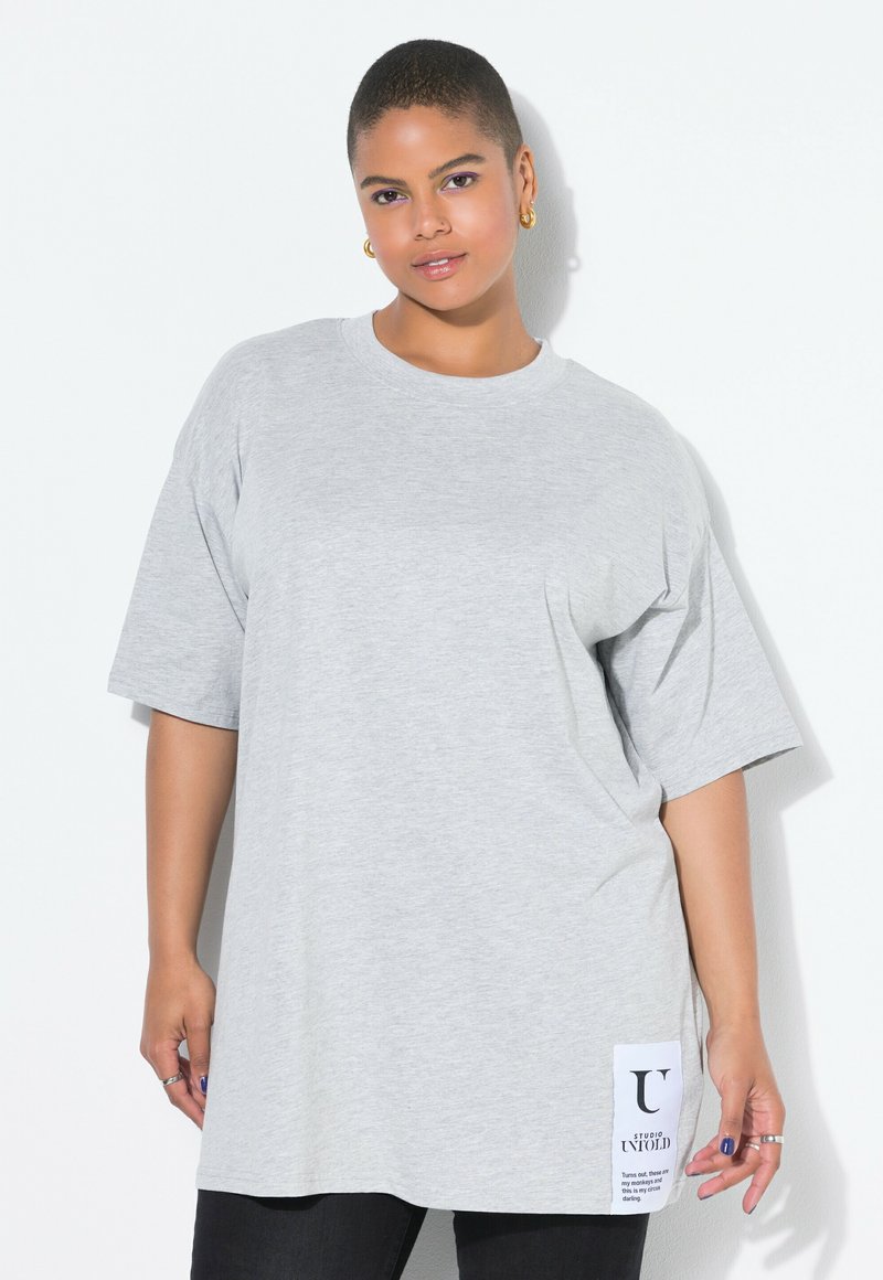 Camiseta de gran tamaño en gris claro con mangas cortas, escote redondo y una etiqueta lateral con texto negro sobre fondo blanco.