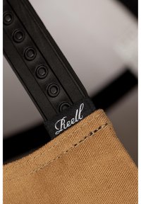 A tan canvas bag with a black adjustable strap features circular hardware and a black label with white lettering reading "Reell."