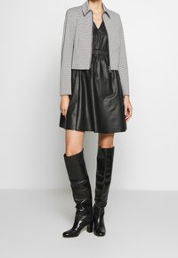 Robe en cuir noir avec une taille élastique, associée à une veste courte à motif pied-de-poule. Bottes noires à talons montantes jusqu'aux genoux complètent la tenue.