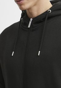 Haut de veste noir zippé fabriqué en tissu texturé, avec un col haut, une fermeture éclair argentée et des accents à cordon.