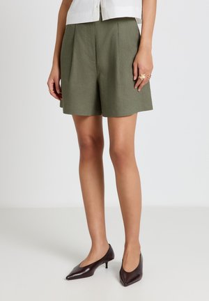 Donna con pantaloncini sartoriali a vita alta verde oliva, camicia bianca, anelli d'oro e décolleté marrone scuro a punta con tacco kitten, in piedi su pavimento bianco.