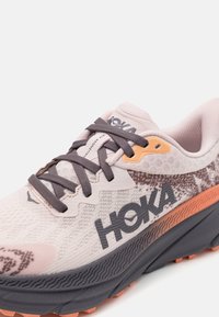 Scarpa da corsa rosa chiaro con superiore testurizzato, lacci scuri e suola ammortizzata. Presenta un logo HOKA e accenti di motivo sottili.