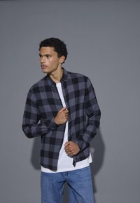 Camicia di flanella a quadri nera e grigia con colletto button-down, maniche lunghe e bottoni davanti, indossata sopra una maglietta bianca.