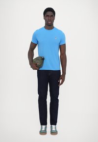 Tricou din bumbac de culoare bleu deschis, cu mâneci scurte, blugi de culoare bleu închis, geantă crossbody de culoare verde și adidași de culoare teal cu șireturi albe și talpă de culoare caramel.