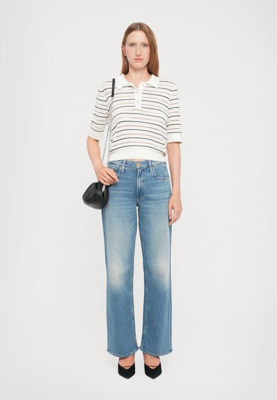 Witte polo met korte mouwen en horizontale strepen, gecombineerd met lichtblauwe wijde jeans en zwarte puntige schoenen. Houdt een kleine zwarte tas vast.