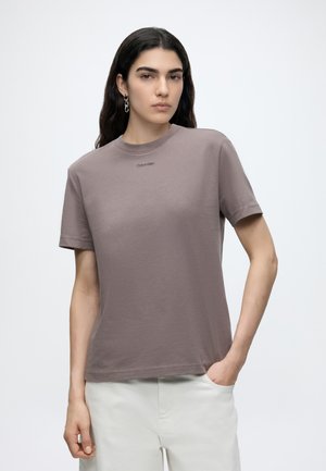 Jeune femme aux longs cheveux foncés porte un t-shirt mauve Calvin Klein et un pantalon blanc, debout avec une main dans la poche devant un fond uni.