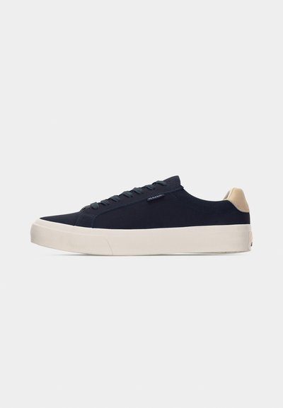 Baskets basses bleu marine avec languette de talon beige, semelle épaisse blanche, et fermeture à lacets, vues de côté gauche sur fond gris clair.