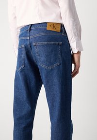 Jeans de mezclilla azul oscuro con una etiqueta de cuero de Calvin Klein en la cintura. Bolsillos traseros estándar con costuras visibles y un corte recto.
