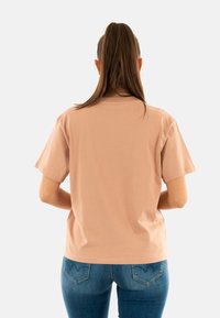 Camiseta de algodón rosa claro con mangas cortas y un corte relajado, mostrada desde la parte de atrás. Combinada con jeans azules que presentan un diseño de bolsillo curvado.
