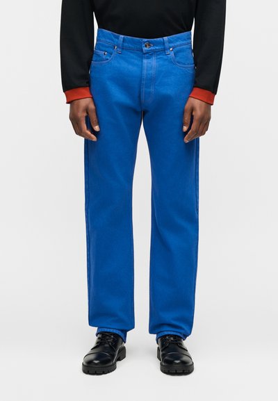 Άνδρας που φοράει φωτεινά μπλε τζιν με ίσιο πόδι, μαύρα παπούτσια, μαύρο πουκάμισο με μακριά μανίκια και κόκκινες μανσέτες, στέκεται μπροστά σε λευκό φόντο.