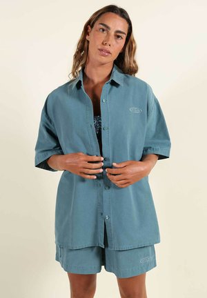 Femme portant une chemise ample à manches courtes bleu sarcelle avec des boutons et un short assorti, debout devant un fond beige uni.