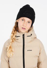 Schwarze gerippte Strickmütze mit geprägtem "SKI & SNOW CLUB"-Text, kombiniert mit einer beigen Steppjacke mit Reißverschluss und verstellbarer Kapuze.