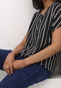 Blouse à rayures noires et blanches avec manches courtes, col en V et tissu doux ; portée avec un jean en denim bleu.