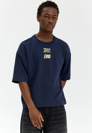 Mladý muž má na sebe tmavomodré oversized tričko so žltým textom "EVERYDAY INC. BASED IN CEST (UTC+2) EVRD" a čierne džínsy, stojí na bielom pozadí.