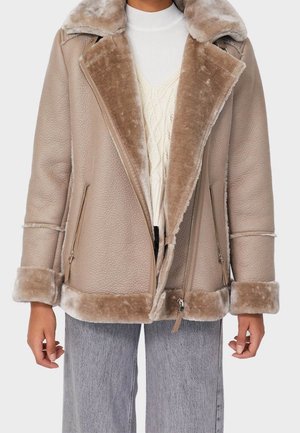 Veste en peau lainée beige avec grand col, fermeture éclair devant et poignets, portée sur un pull torsadé crème et un jean gris.