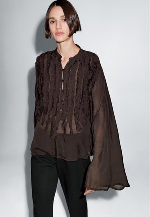 Zwarte doorschijnende blouse met ruches en een knoopsluiting. Voorzien van lange, brede mouwen en een losse pasvorm, gecombineerd met zwarte broek.