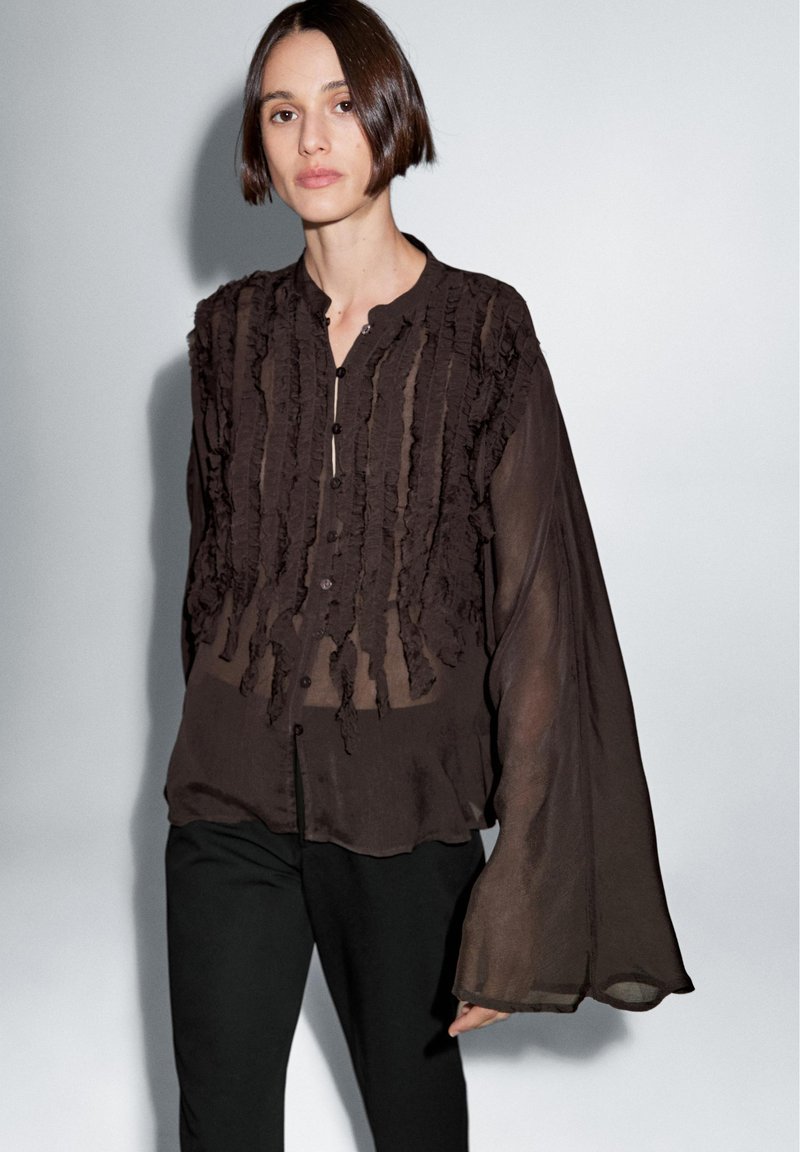 Sort transparent bluse med flæsedetaljer og knaplukning. Har lange, vide ærmer og en afslappet pasform, kombineret med sorte bukser.