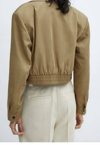 Abgeschnittene, braune Bomberjacke mit glatter Textur, elastischem Bund und geknöpften Ärmelbündchen. Trägt man über hellen Hosen.