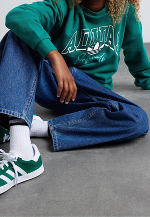 Grüner Pullover mit weißem Text und Logo, hochtaillierte blaue Jeans, weiße und grüne Sneakers sowie weiße Socken mit schwarzen Streifen.