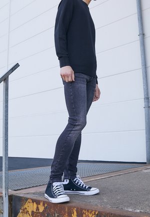 Osoba stoi na zewnątrz na zardzewiałej metalowej platformie, ubrana w czarne trampki typu high-top, ciemne obcisłe jeansy i czarną bluzkę z długim rękawem.