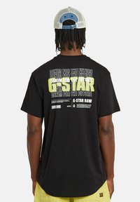 Camiseta de algodón negra con texto en blanco y amarillo neón en la parte posterior. Cuello redondo y mangas cortas. Etiqueta visible en el lado.