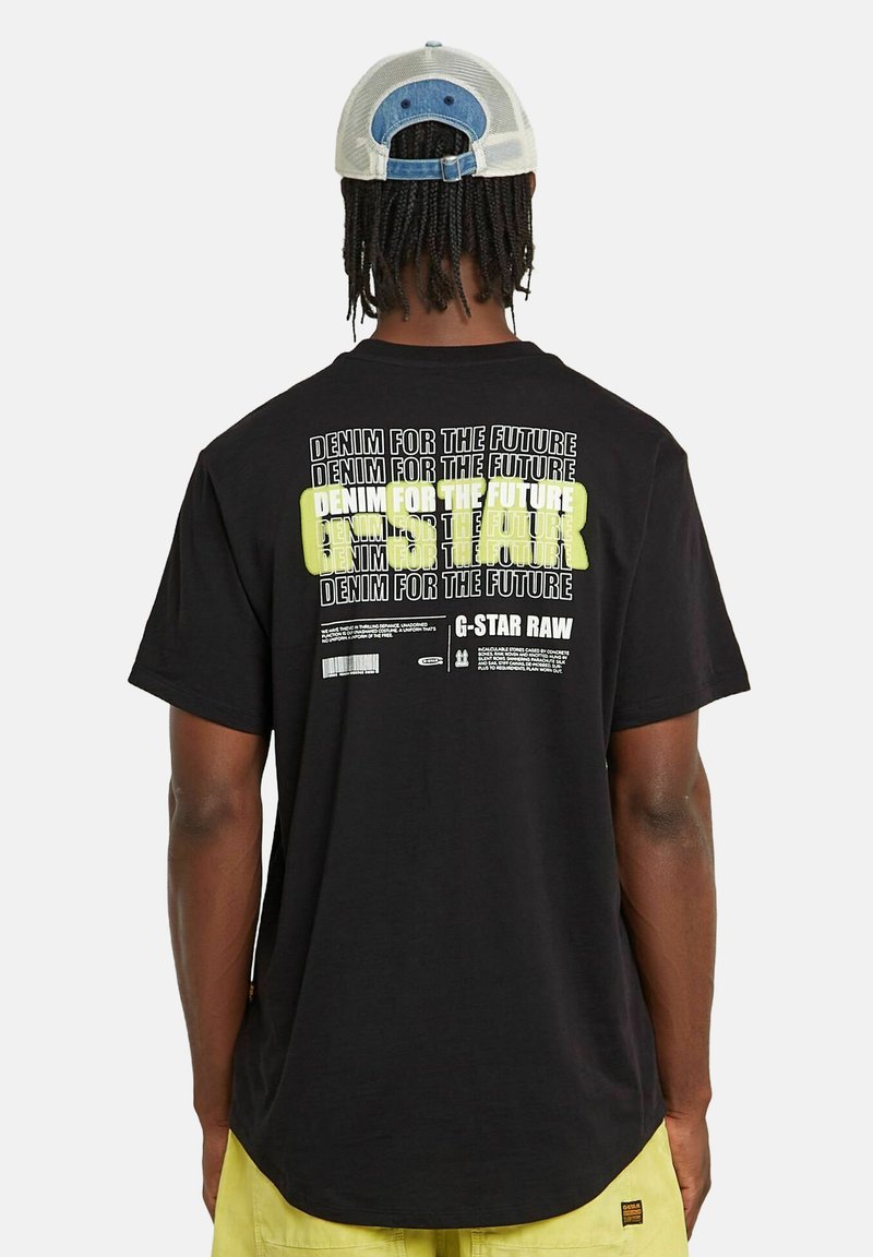 Camiseta de algodón negra con texto en blanco y amarillo neón en la parte posterior. Cuello redondo y mangas cortas. Etiqueta visible en el lado.